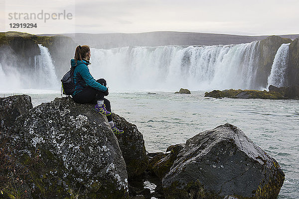 Porträt einer Wanderin  die auf einem Felsen sitzt und den plätschernden Godafoss-Wasserfall betrachtet  Island