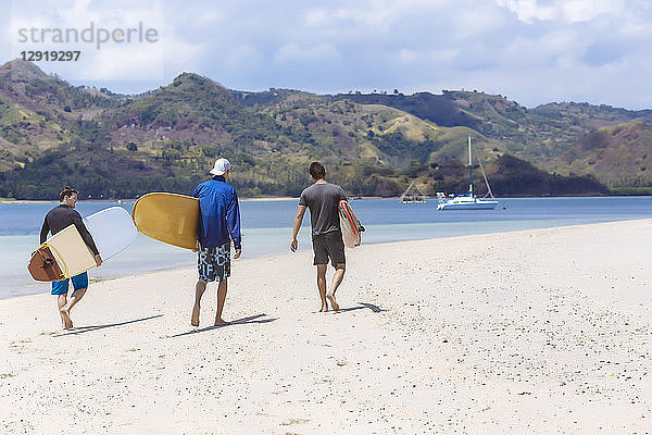 Rückansicht von drei männlichen Surfern  die am Strand spazieren gehen