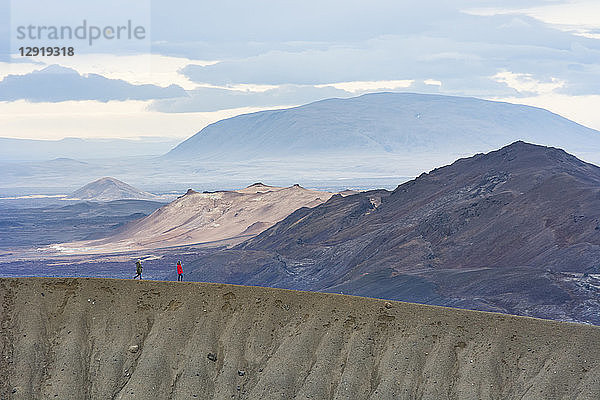 Zwei Personen in farbenfroher Kleidung spazieren entlang des Kammes des Viti-Kraters  mit einer bergigen Landschaft im Hintergrund  im Vulkangebiet Krafla  in der Nähe des Myvatn-Sees in Nordisland.