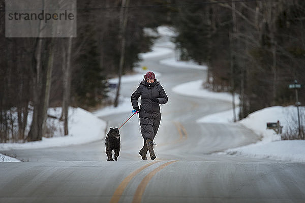Vorderansicht einer reifen Frau  die mit ihrem Hund auf einer Straße im Wald spazieren geht  im Winter