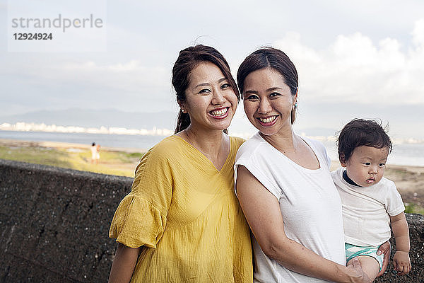 Porträt von zwei Japanerinnen  eine mit einem Kleinkind  die auf einer Promenade am Meer stehen und in die Kamera lächeln.