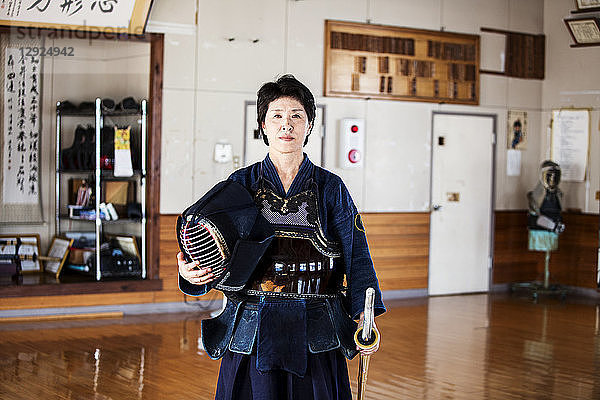 Japanische Kendo-Kämpferin steht in einer Turnhalle  hält Kendo-Maske und Schwert und schaut in die Kamera.