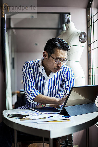 Japanischer männlicher Modedesigner  der in seinem Atelier arbeitet  am Tisch sitzt und auf ein digitales Tablett schaut.