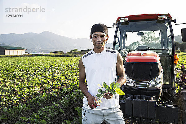 Ein japanischer Bauer steht vor einem roten Traktor in einem Sojabohnenfeld und schaut in die Kamera.