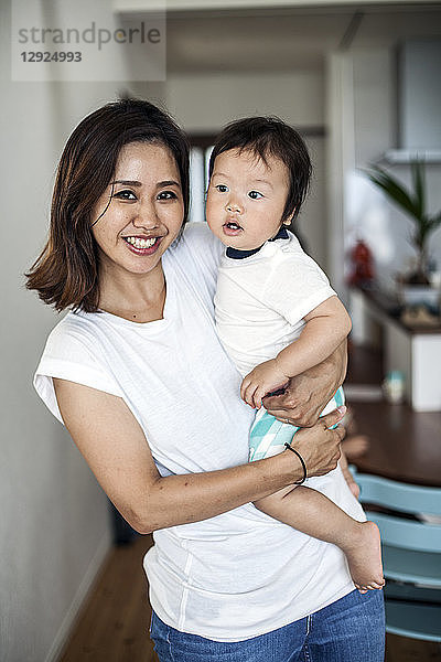Porträt einer Japanerin  die in einem Wohnzimmer steht und ein Kleinkind trägt und in die Kamera lächelt.