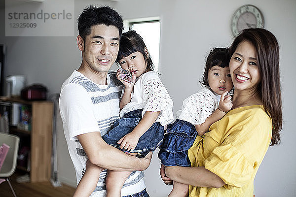 Porträt eines japanischen Mannes und einer japanischen Frau mit zwei jungen Mädchen  die in einem Wohnzimmer stehen und in die Kamera lächeln.