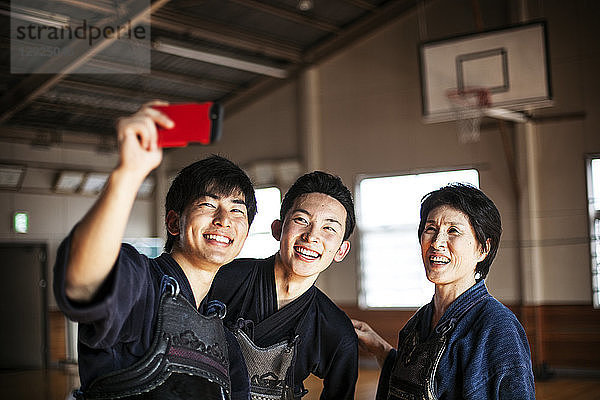 Lächelnde weibliche und zwei männliche japanische Kendo-Kämpfer stehen in einer Turnhalle und nehmen Selfie mit dem Handy ein.