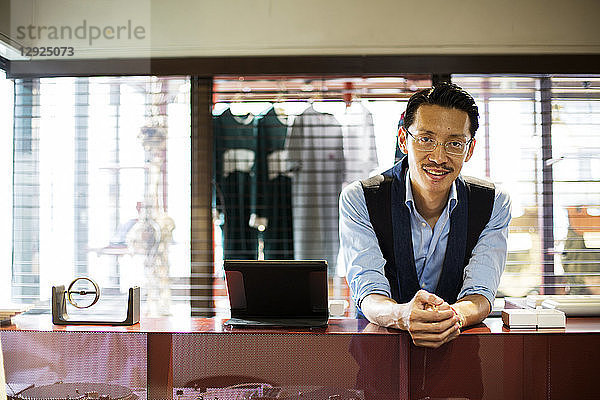 Japanischer Verkäufer mit Schnurrbart und Brille steht in einem Bekleidungsgeschäft an der Theke und lächelt in die Kamera.
