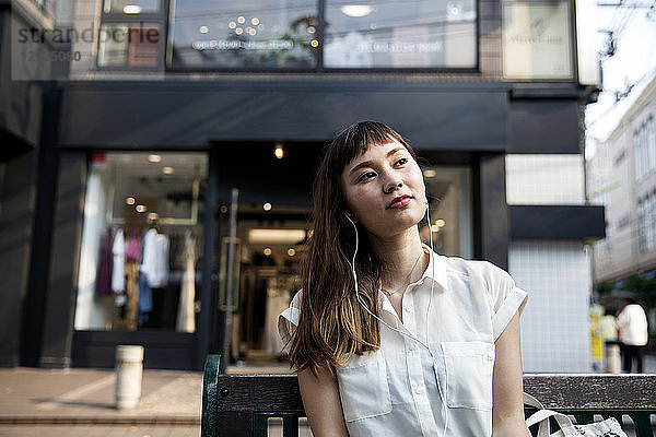 Japanerin mit langen braunen Haaren in weißer  kurzärmeliger Bluse  die auf einer Bank sitzt  Kopfhörer trägt und Musik hört.