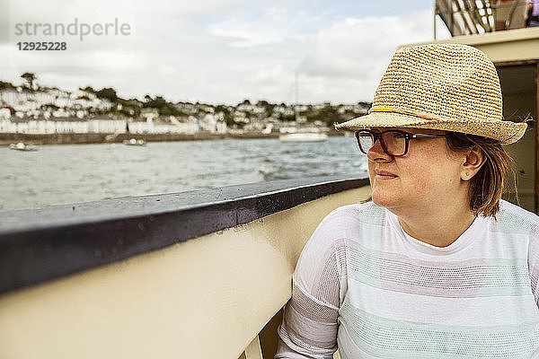 Eine Frau mit Strohhut auf einem Boot  die an einem bedeckten Tag an der Küste auf die Stadt hinaussieht.