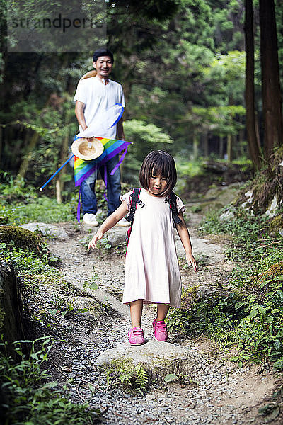Im Wald stehendes japanisches Mädchen in hellrosa Sonnenkleid und mit Rucksack  im Hintergrund ein Mann.
