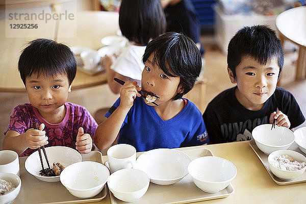 Schrägansicht von drei Jungen  die in einer japanischen Vorschule an einem Tisch sitzen und mit Stäbchen essen.