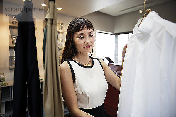 Lächelnde japanische Verkäuferin steht im Bekleidungsgeschäft und schaut auf das Hemd.
