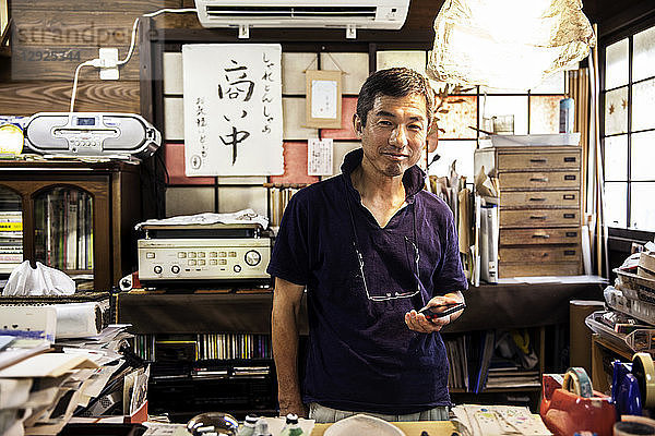 Japanischer Mann steht in einer Washi-Produktionswerkstatt  hält ein Smartphone in der Hand und lächelt in die Kamera.