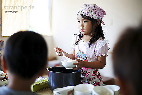 Kopftuch tragendes Mädchen  das in einer japanischen Vorschule an einem Tisch steht und den Kindern das Mittagessen serviert.