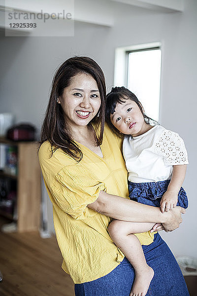 Porträt einer Japanerin  die in einem Wohnzimmer steht und ein junges Mädchen trägt  das in die Kamera lächelt.