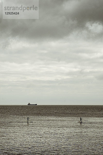 Blick von den Klippen aufs Meer  zwei Personen paddeln auf ruhiger See an Bord  und am Horizont ein großes Frachttankschiff.
