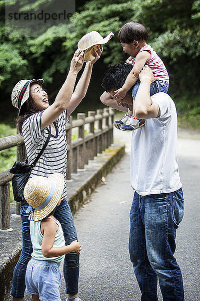 Japanisches Mädchen  lächelnde Frau mit Hut und Mann  der ein Kleinkind auf den Schultern trägt und auf einer Holzbrücke steht.