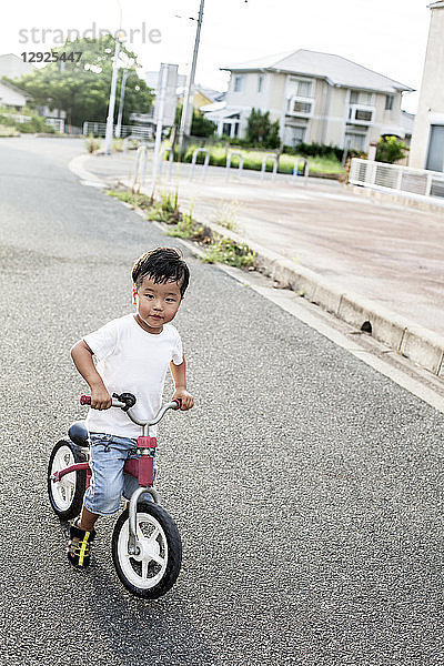 Porträt eines japanischen Jungen  der mit einem Fahrrad auf der Straße spielt und in die Kamera schaut.