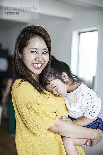 Porträt einer Japanerin  die in einem Wohnzimmer steht und ein junges Mädchen trägt  das in die Kamera lächelt.