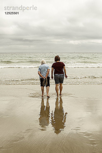 Rückansicht einer grauhaarigen älteren Frau und einer jüngeren Frau  die mit ausgezogenen Schuhen in flachen Wellen auf einem Sandstrand paddeln.
