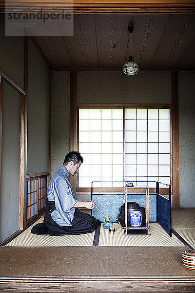 Japanischer Mann in traditionellem Kimono  der während einer Teezeremonie auf dem Boden kniet und einen Hishaku  eine Bambusschöpfkelle  benutzt.