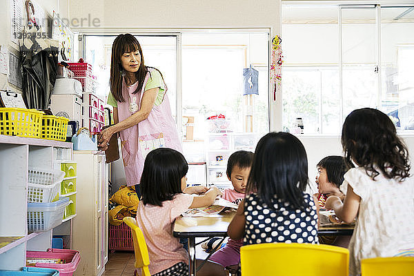 Eine junge Lehrerin und eine Gruppe von Kindern in einer japanischen Vorschule an einem Tisch sitzend.