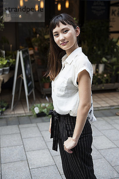 Japanerin mit langen braunen Haaren und weißer  kurzärmeliger Bluse  steht in einer Straße und lächelt in die Kamera.