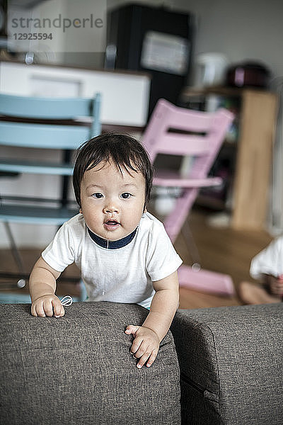 Porträt eines japanischen Kleinkindes  das auf einem grauen Sofa steht und neugierig in die Kamera schaut.