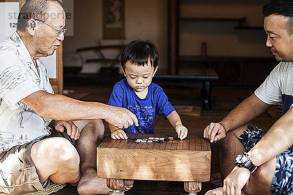 Zwei japanische Männer und ein kleiner Junge sitzen auf dem Boden auf der Veranda eines traditionellen japanischen Hauses und spielen Go.