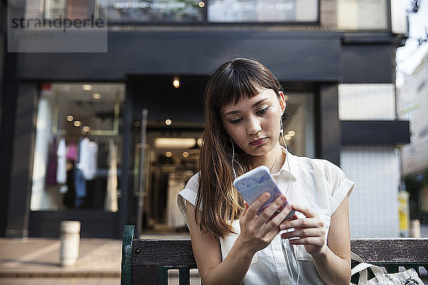 Japanerin mit langen braunen Haaren und weißer  kurzärmeliger Bluse  steht in einer Straße und schaut auf ein Mobiltelefon.