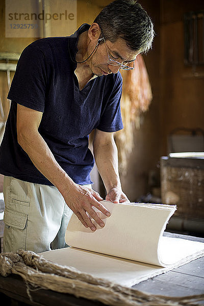 Japanischer Mann  der in einer Werkstatt steht und Blätter von traditionellem Washi-Papier durchsieht.