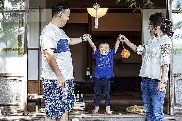 Japanischer Mann und Frau stehen auf der Veranda eines traditionellen japanischen Hauses  spielen mit einem kleinen Jungen und halten sich an den Händen.