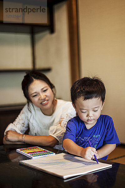 Lächelnde japanische Frau und kleiner Junge sitzen an einem Tisch und zeichnen mit Farbstiften auf weißes Papier.