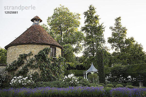 Blick auf den kleinen runden Steinturm und den Pavillon von einem ummauerten Garten mit Bäumen und Blumenbeeten aus.