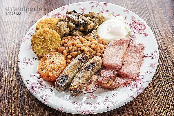 Traditionelles englisches Frühstück mit Speck  Würstchen  gegrillten Tomaten und Champignons  gebackenen Bohnen  Kartoffeln  Hash Browns  Kuchen und Ei auf einem Holztisch