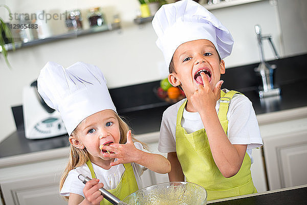 Zwei junge Kinder glücklich Kinder Junge und Mädchen Familie mit Schürze und Kochmütze Vorbereitung lustige Cookies in der Küche zu Hause.
