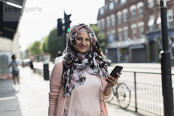 Porträt einer lächelnden  selbstbewussten Frau mit Smartphone  die einen geblümten Hijab auf einem städtischen Bürgersteig trägt