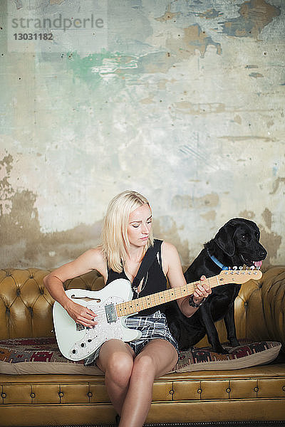 Junge Frau mit Hund spielt E-Gitarre auf Sofa
