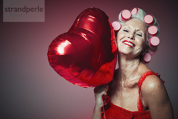 Porträt einer verspielten älteren Frau mit Lockenwicklern  die einen Luftballon in Herzform hält