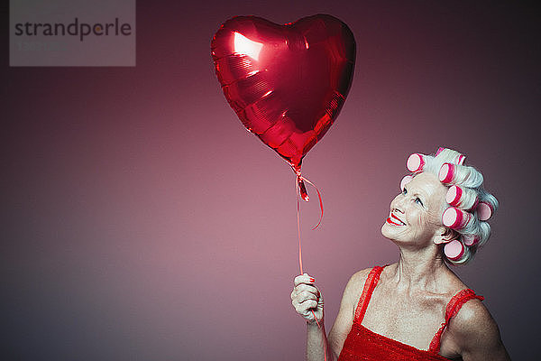 Porträt einer lächelnden älteren Frau mit Lockenwicklern  die einen Luftballon in Herzform hält