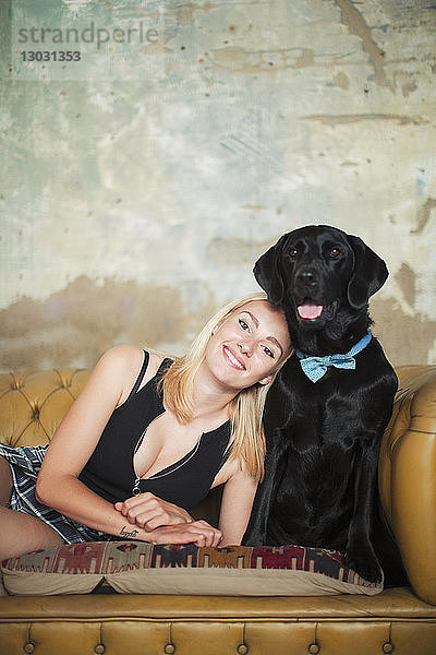 Porträt einer lächelnden  unbekümmerten jungen Frau mit schwarzem Hund und Fliege auf dem Sofa