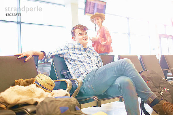 Junger Mann sitzt am Flughafen  Rucksack neben ihm  junge Frau steht neben ihm und schaut zu ihm