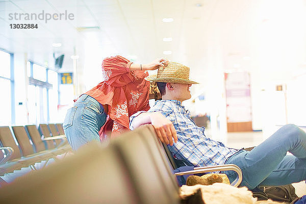 Junger Mann sitzt am Flughafen  junge Frau setzt ihm ihren Hut auf