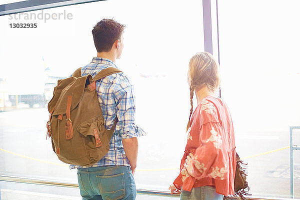 Junges Paar am Flughafen  Rucksäcke tragend  aus dem Fenster schauend