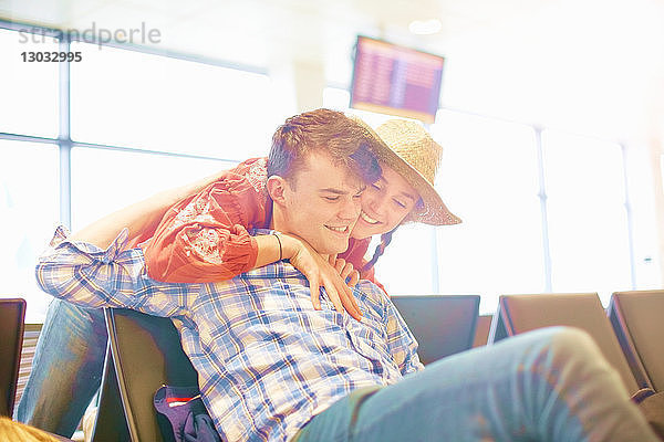 Junger Mann sitzt am Flughafen  junge Frau streckt sich über den Sitz  um ihn zu umarmen