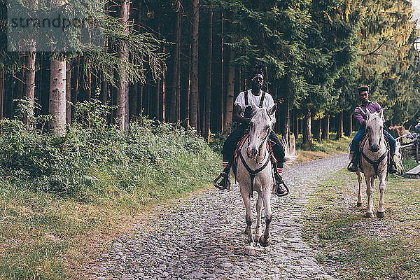 Zwei junge Männer reiten auf Pferden auf ländlicher Kopfsteinpflasterstraße  Primaluna  Trentino-Südtirol  Italien
