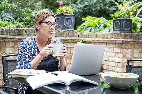 Schwangere Frau im mittleren Erwachsenenalter schaut auf den Laptop auf dem Terrassentisch