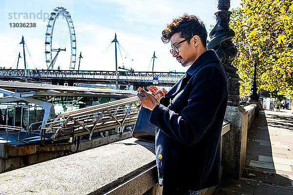 Geschäftsmann mit Smartphone am Embankment  London Eye und Hungerford Bridge im Hintergrund  London  Großbritannien