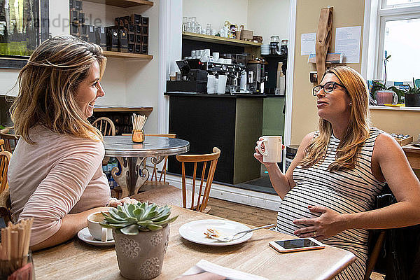 Schwangere Frau im mittleren Erwachsenenalter und beste Freundin unterhalten sich bei einem Kaffee im Café